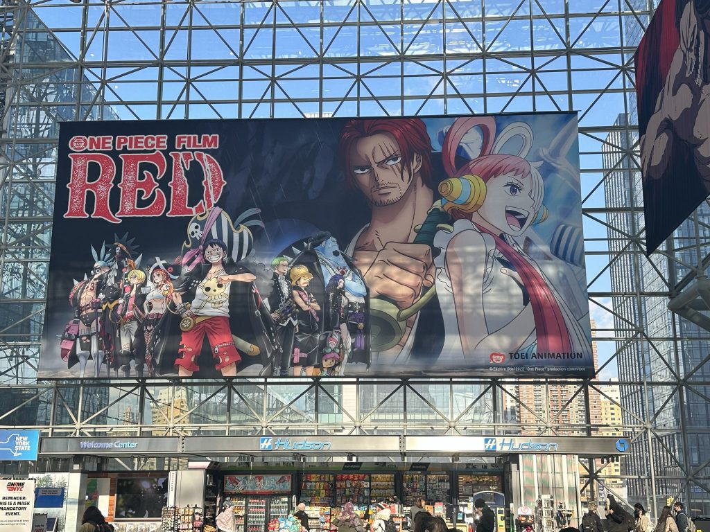 Banner of One Piece Film Red head up in front of glass walls of the Jacob K. Javits Convention Center. It features the characters Uta, Shanks, and the Straw Hat Pirates wearing punk rock inspired pirate-y outfits.